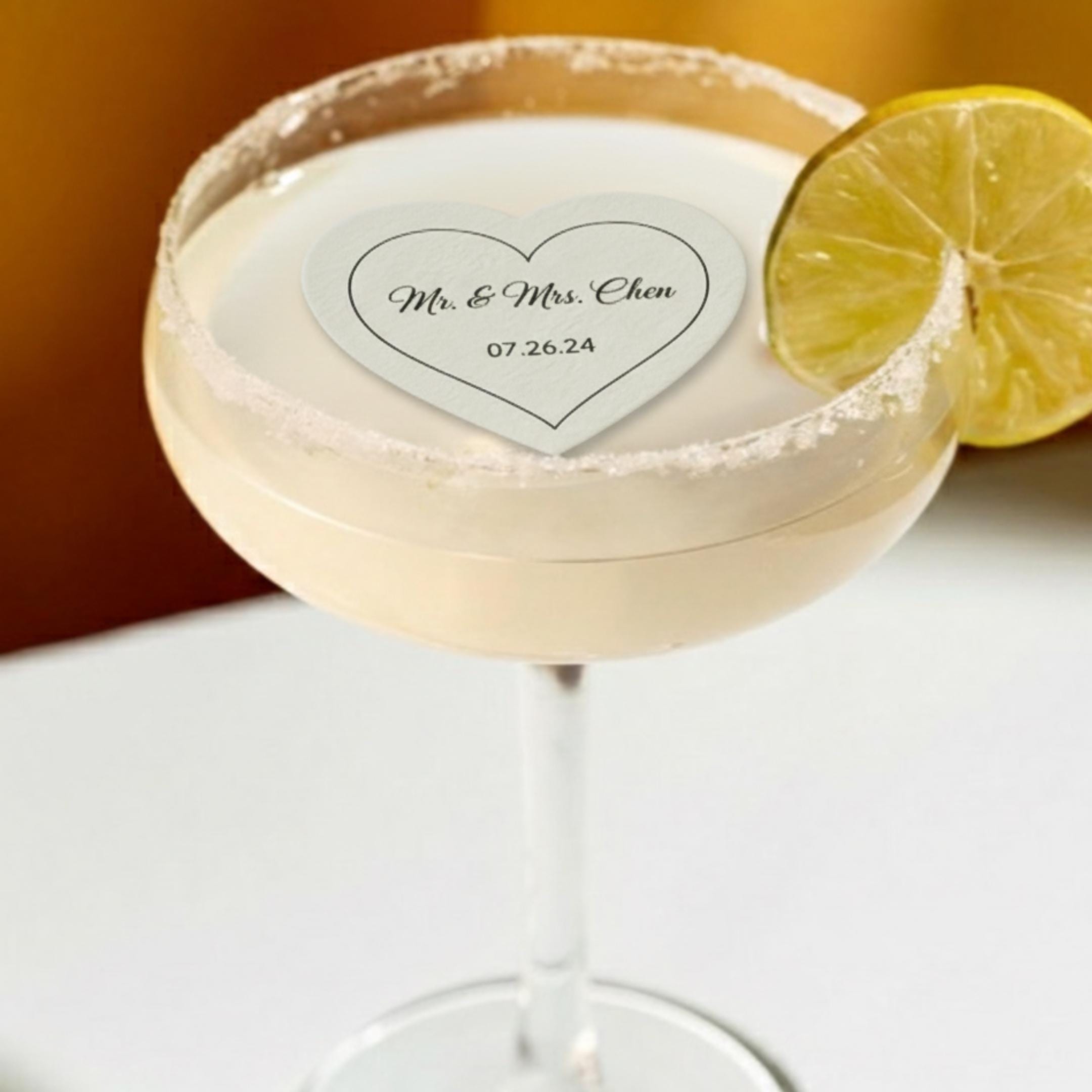 A cocktail in a stemmed glass with a sugared rim, garnished with a lemon slice. A heart-shaped paper with the text "Mr. & Mrs. Chen 07.26.24" floats in the drink.