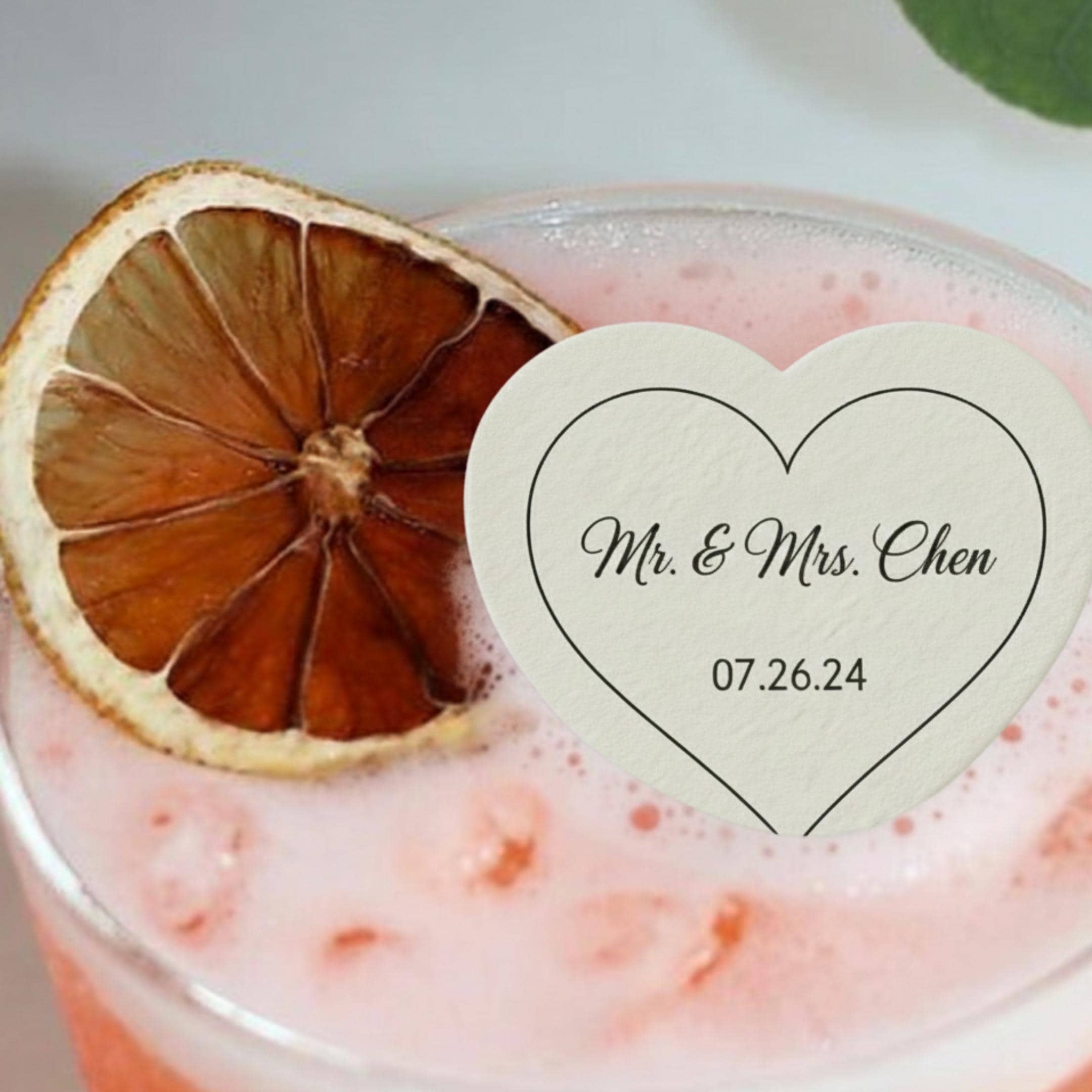 A cocktail with a dried orange slice garnish and a heart-shaped coaster. The coaster has the text "Mr. & Mrs. Chen" and the date "07.26.24" in black script. The drink is pink and bubbly.