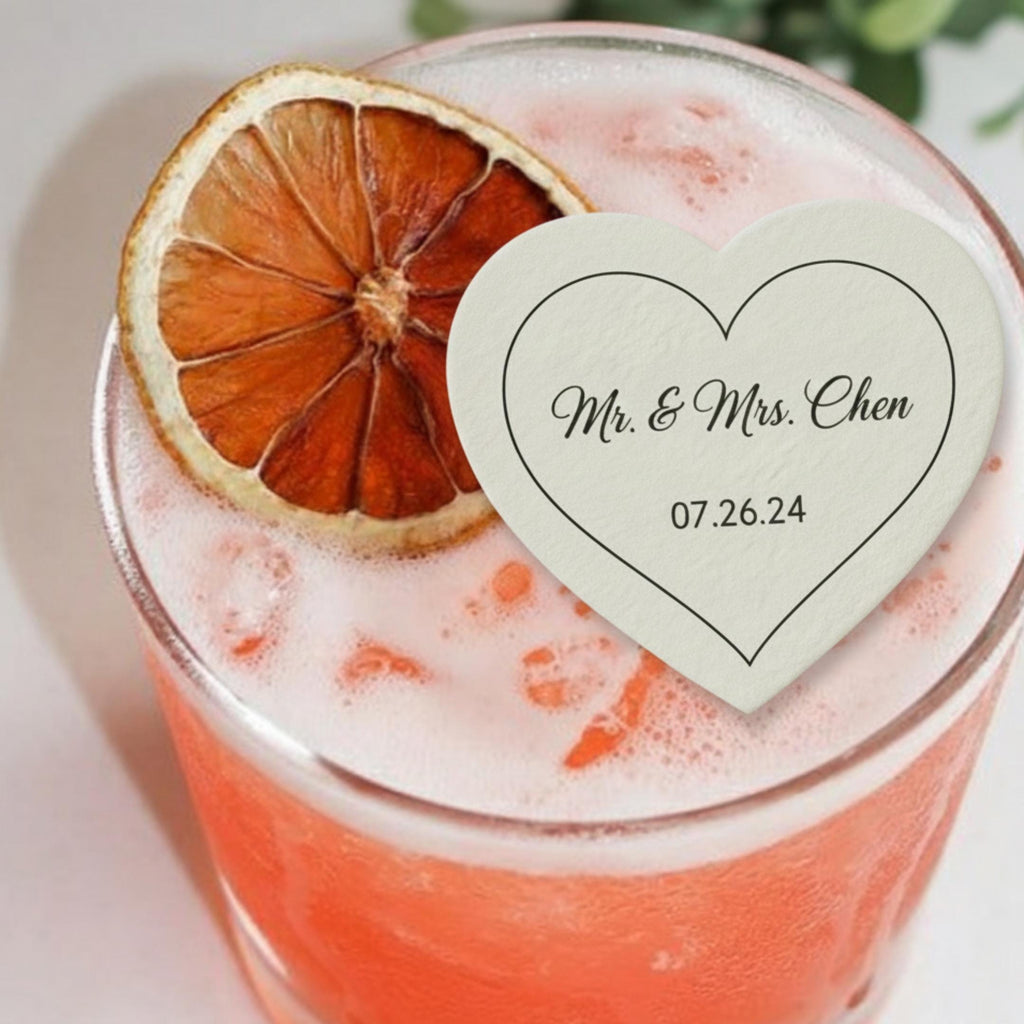 A cocktail in a glass, garnished with a dried orange slice. A heart-shaped coaster with the text "Mr. & Mrs. Chen" and the date "07.26.24" rests on the drink. The drink is a vibrant orange color.