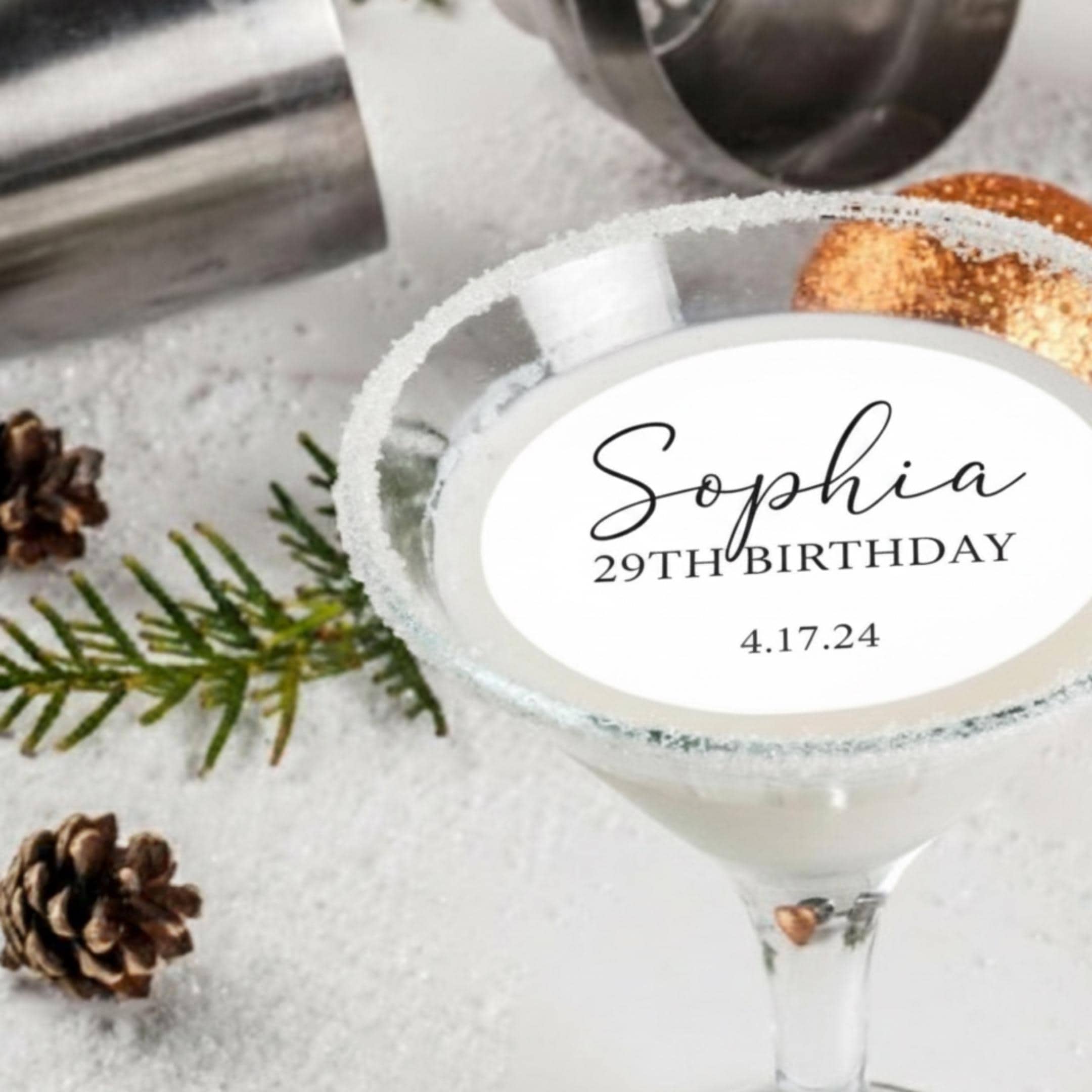 A martini glass with a sugar-rimmed edge, filled with a white cocktail. The glass has a white oval sticker with the name "Sophia", "29TH BIRTHDAY", and the date "4.17.24". A stainless steel cocktail shaker and pine cones are in the background.