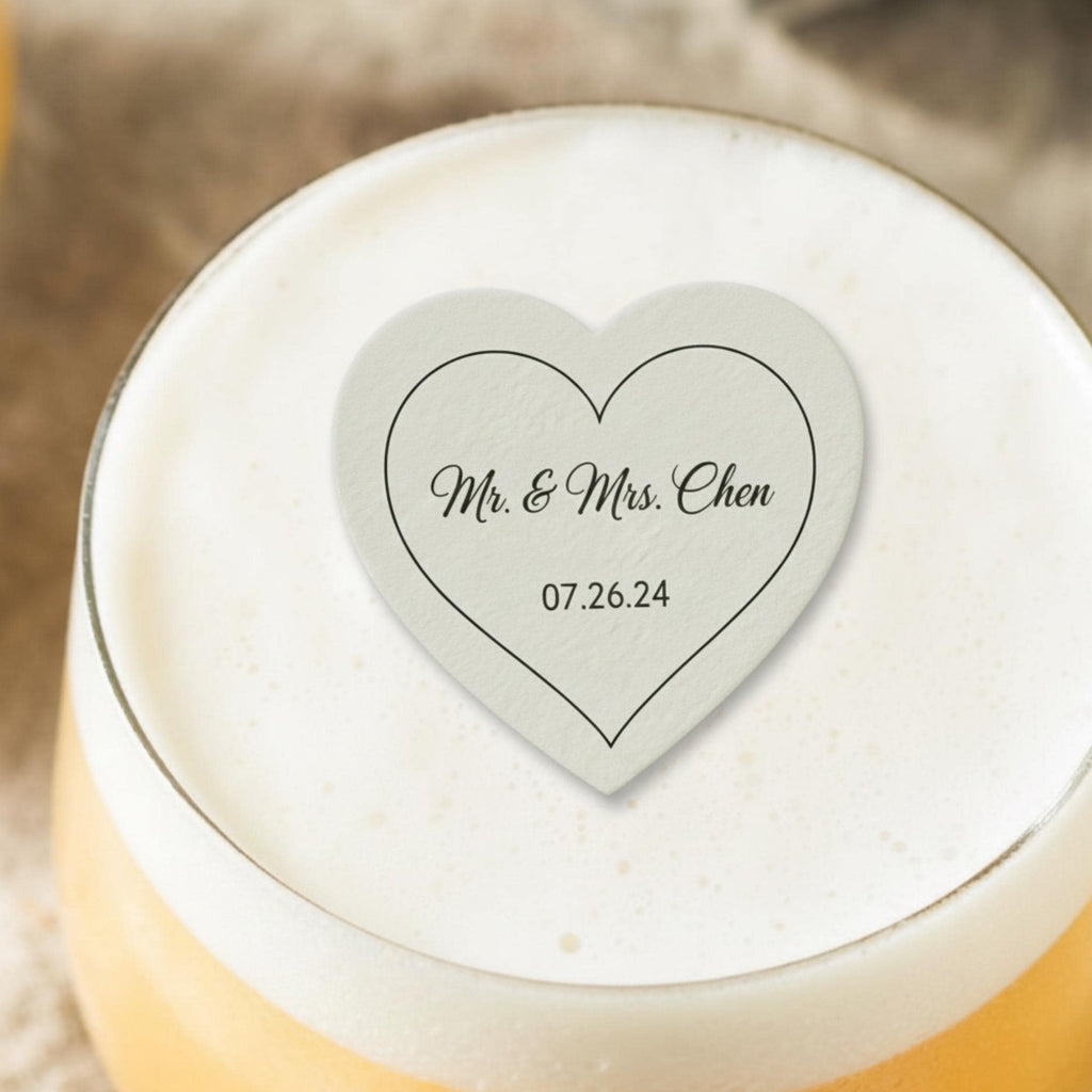 A heart-shaped coaster with the text "Mr. & Mrs. Chen" and the date "07.26.24" printed in black. The coaster is light gray and sits atop a glass of beer with a foamy head. The image is a close-up shot.