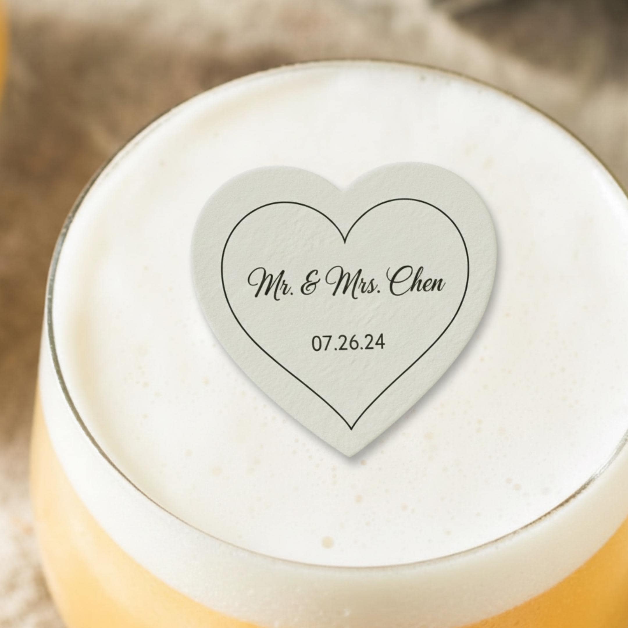 A heart-shaped coaster with the text "Mr. & Mrs. Chen" and the date "07.26.24" printed in black. The coaster is light gray and sits atop a glass of beer with a foamy head. The image is a close-up shot.