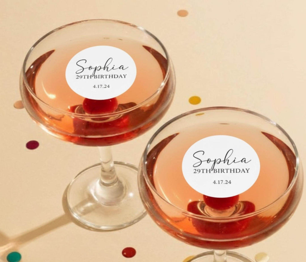 Two champagne coupe glasses filled with a pink beverage, each with a white circular sticker that reads "Sophia 29th Birthday 4.17.24". The glasses sit on a light beige surface with scattered confetti.