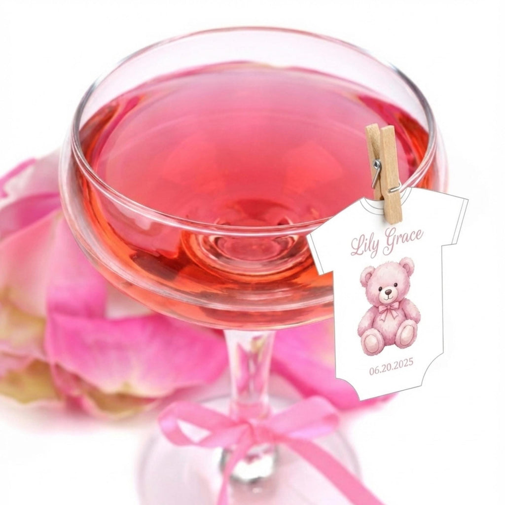 Baby Shower Drink Toppers A champagne glass filled with pink liquid, possibly rosé, with a decorative baby onesie attached with a clothespin. The onesie features a pink teddy bear and the name "Lily Grace" with the date 06.20.2025.