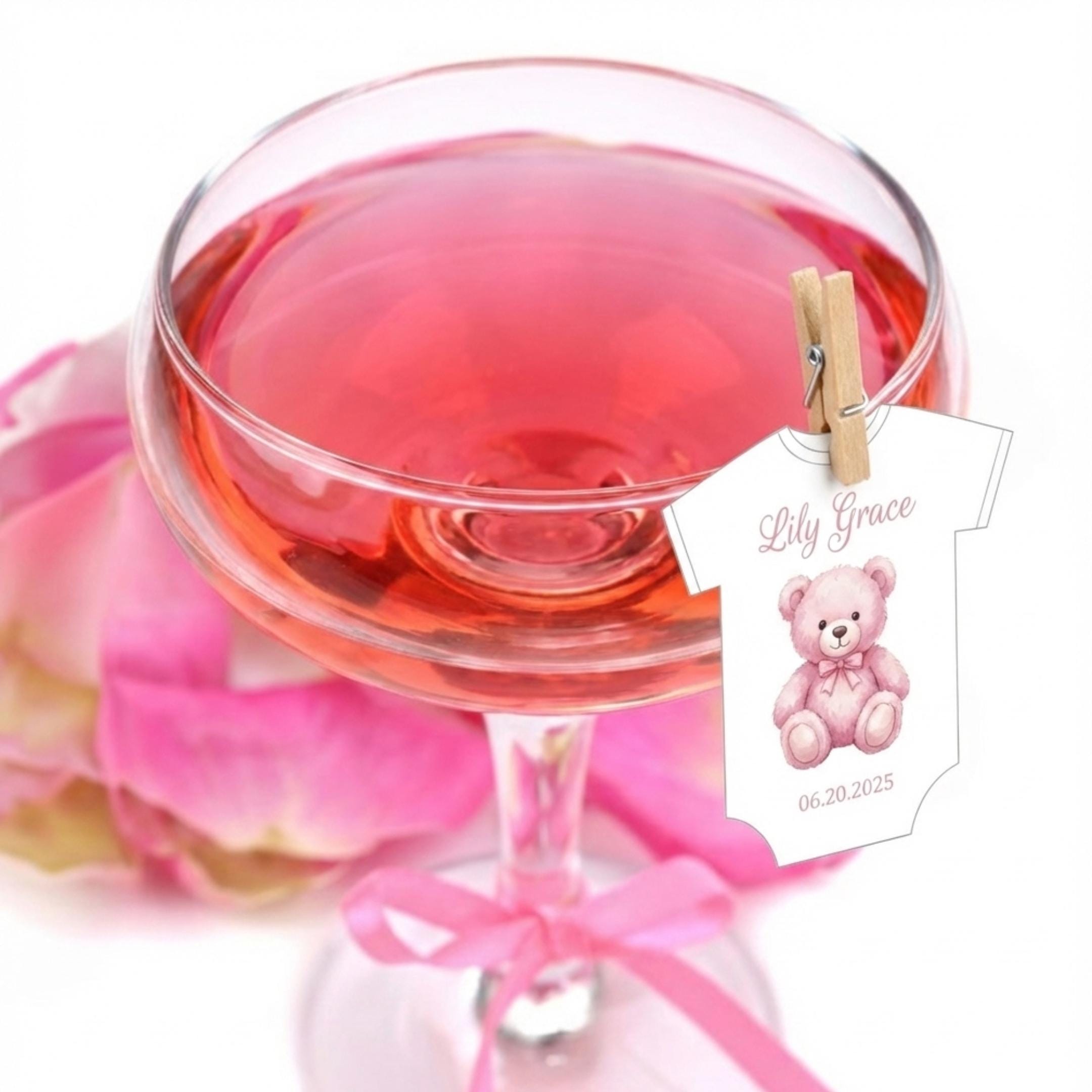 Baby Shower Drink Toppers A champagne glass filled with pink liquid, possibly rosé, with a decorative baby onesie attached with a clothespin. The onesie features a pink teddy bear and the name "Lily Grace" with the date 06.20.2025.