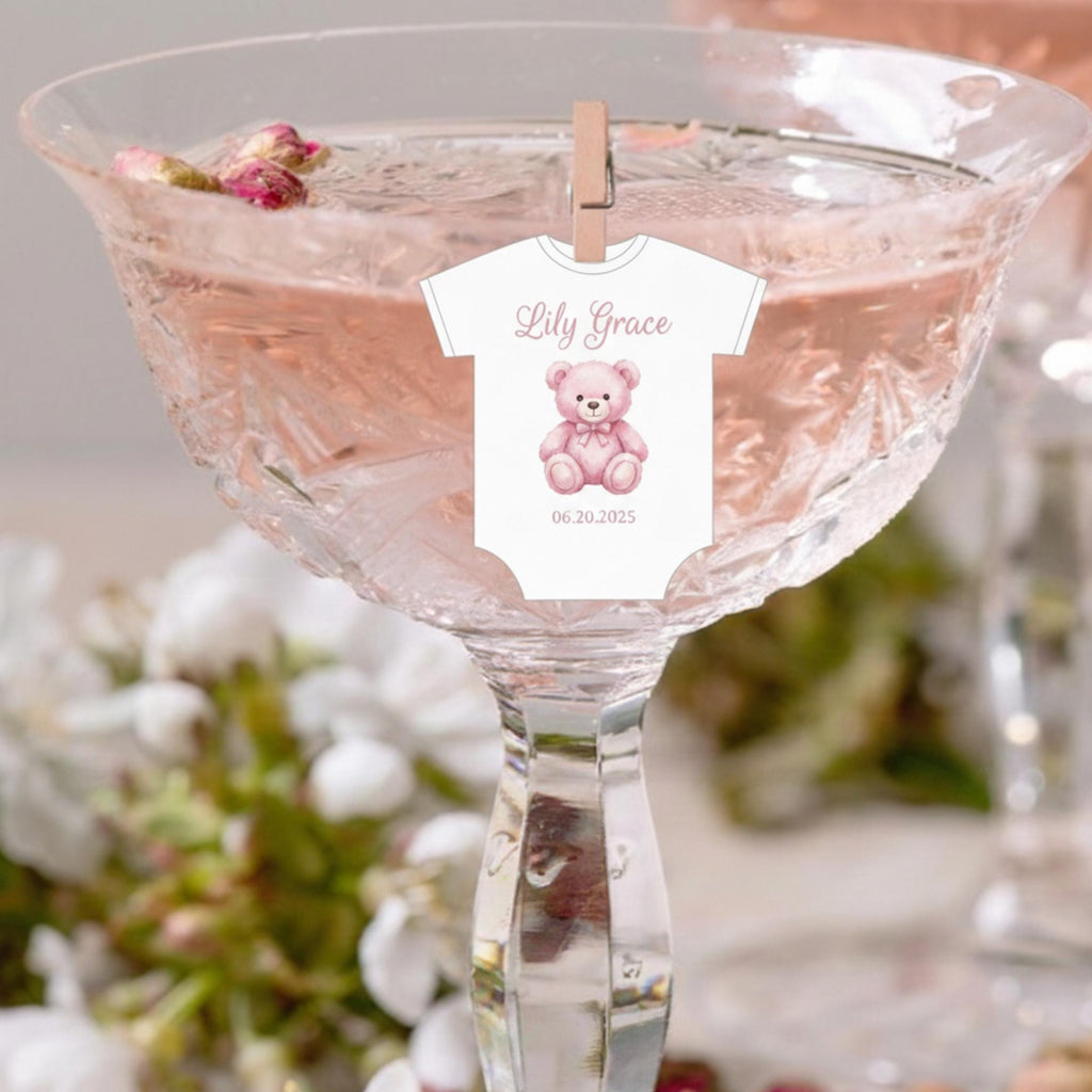 Baby shower edible drink toppers in cute onesie shape made from wafer paper, designed to float on coffee, milk foam, mocktails, and party drinks. These precut toppers are printed with food-safe edible ink, adding a soft and charming touch to baby shower, gender reveal, and welcome baby celebrations. Perfect for themed party decor, they create a stylish and photo-ready presentation while enhancing the overall event setup A white baby onesie with a pink teddy bear graphic